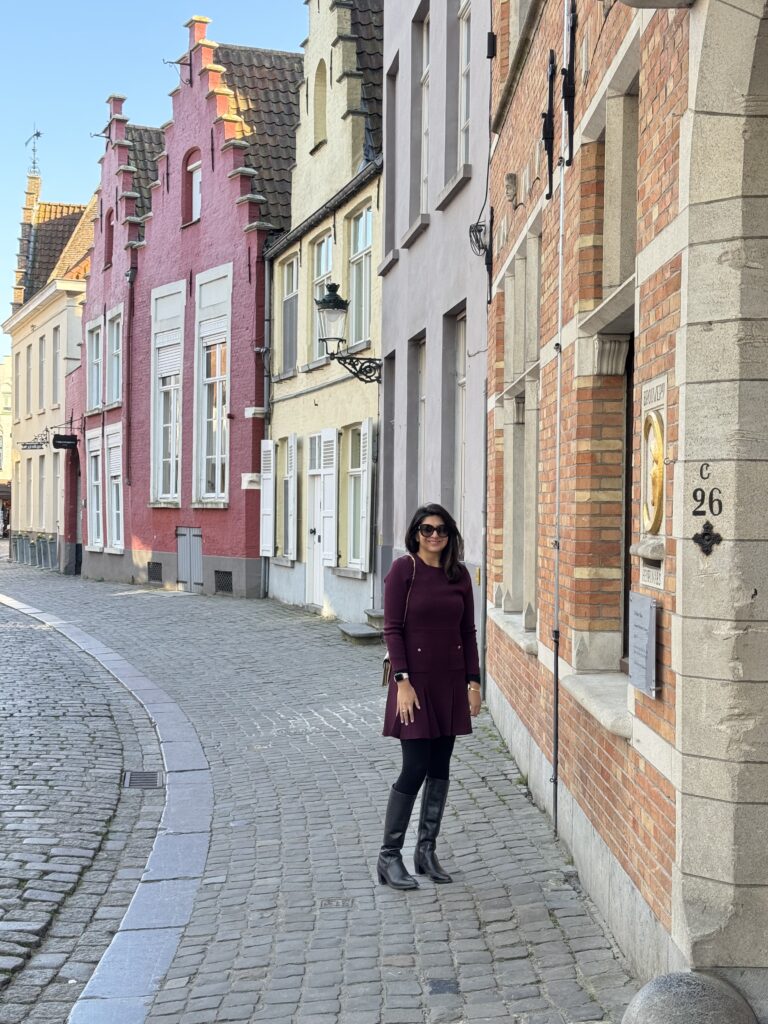 A woman in a red coat stands on a cobblestone street, surrounded by historic buildings and a clear blue sky.
