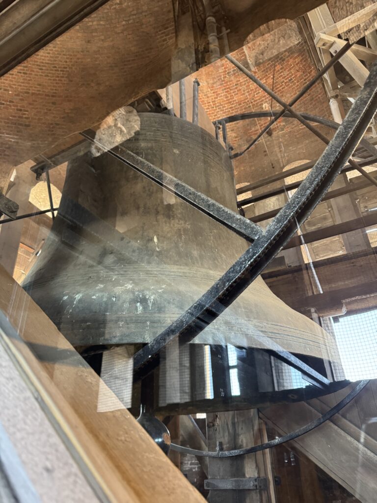 A large bell hangs inside a building, illuminated by light streaming through a nearby window.